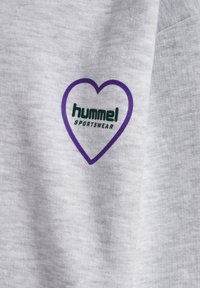 Le vêtement de sport en tissu gris présente un contour de cœur violet avec "hummel" et "SPORTSWEAR" imprimés en vert. Texture douce, design décontracté.