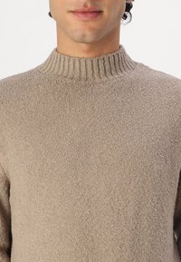 Mann in einem beigen, strukturierten Strickpullover mit einem gerippten Stehkragen, zu sehen von den Schultern bis zum Kinn vor einem schlichten Hintergrund.