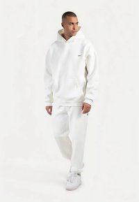 Weißer Kapuzenpullover mit Kängurutasche und Logo auf der Brust, kombiniert mit passenden weißen Jogginghosen und weißen Sneakern. Glatte Textur.