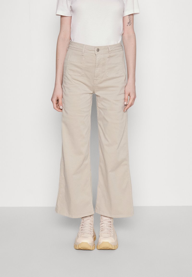Pantaloni beige a gamba larga con tasche frontali, chiusura a bottone e orlo leggermente sfilacciato, abbinati a sneakers beige leggere con suola dorata.