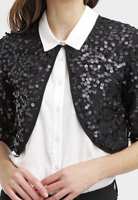 Černá krátká bunda pokrytá kulatými černými a stříbrnými flitry, pod níž je bílá košile s límečkem. Měkká látka s přiléhavým střihem.