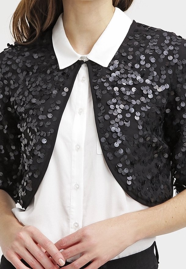 Černá krátká bunda pokrytá kulatými černými a stříbrnými flitry, pod níž je bílá košile s límečkem. Měkká látka s přiléhavým střihem.