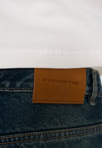 Cămașă albă băgată într-o pereche de blugi albaștri de denim, cu o etichetă maro din piele inscripționată cu „PROHIBITED” la talie.