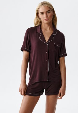 Kobieta ubrana w ciemnofioletowy zestaw piżamy z krótkim rękawem na guziki oraz szorty z białym lamówką na jednolitym tle.