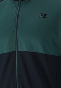 Sportjacka i mörkgrön och marinblå, med dragkedjestängning, slät textur och en liten svart "FZ"-logotyp på bröstet.