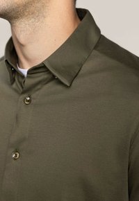 Camicia verde oliva con bottoni e colletto, realizzata in un tessuto morbido e liscio, con bottoni metallici ben in vista.