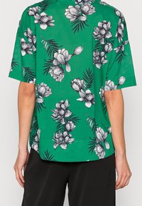 Camisa de manga corta en tela verde con un estampado floral que presenta flores en blanco y negro, complementadas con detalles de hojas.