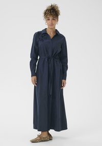 Robe longue bleu marine avec un col, taille ceinturée et détail de broderie florale sur l'épaule gauche. Les matériaux semblent lisses et fluides.