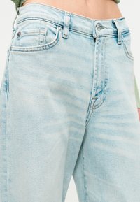 Ανοιχτό μπλε τζιν παντελόνι φορεμένο από ένα άτομο με πράσινη πλεκτή μπλούζα, που δείχνει τις μπροστινές τσέπες, το κουμπί και τις θηλιές της ζώνης.