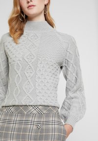Pull en tricot gris clair avec un col montant, présentant des motifs texturés et un design en torsades. Manches longues, associé à un pantalon à carreaux.