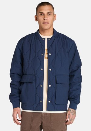 Marineblauer gesteppter Parka mit Druckknöpfen, zwei Fronttaschen, gerippten Bündchen und rundem Ausschnitt, getragen über einem beigen Pullover.
