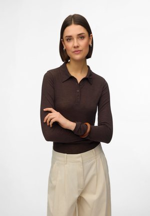 Kvinde med kort mørkt hår iført en mørkebrun langærmet skjorte med krave og beige bukser med høj talje, med armene krydset, neutralt ansigtsudtryk.