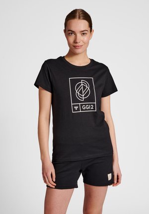 Młoda kobieta w czarnej koszulce z białym geometrycznym wzorem i napisem "GGI2", w połączeniu z czarnymi szortami, stojąca na jednolitym tle.