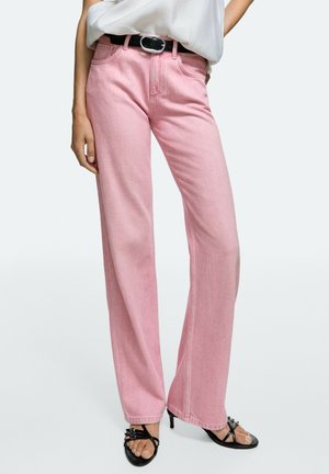 Person trägt hoch taillierte pinke Jeans, einen schwarzen Gürtel, ein weißes eingestecktes Oberteil und schwarze Sandalen mit Absatz, steht vor einem schlichten Hintergrund.