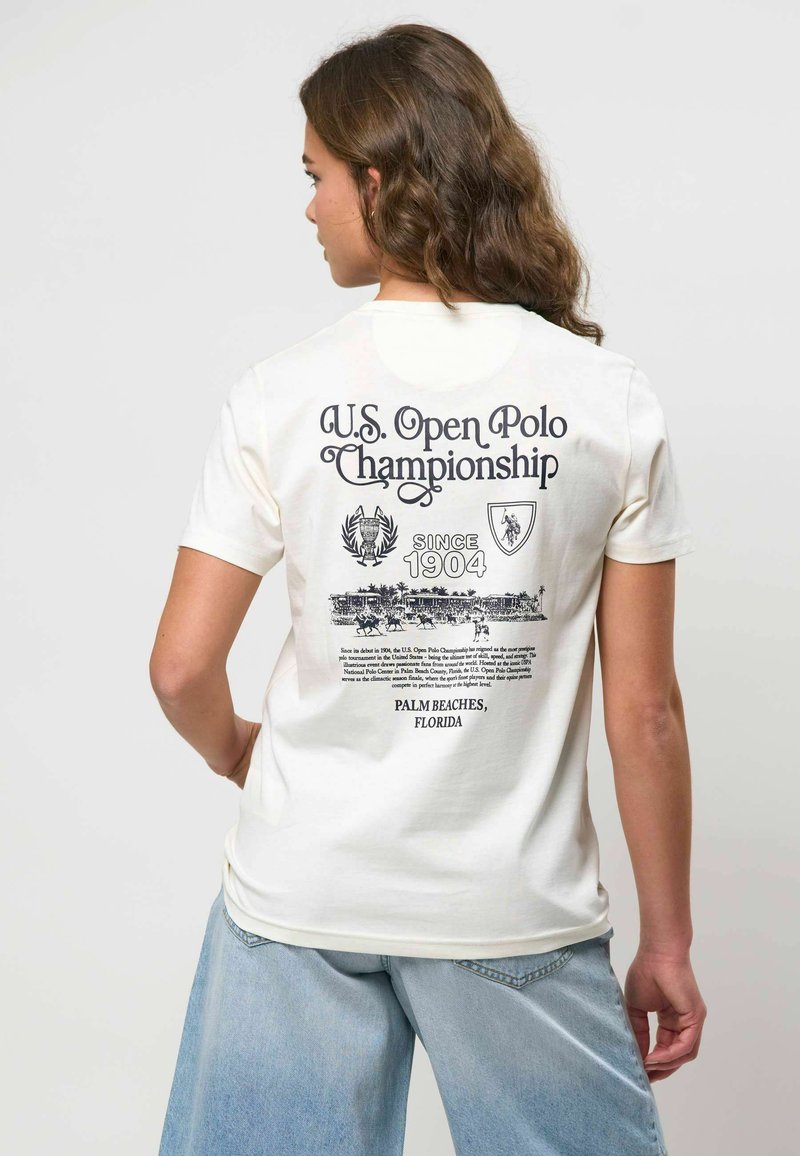 Femme aux longs cheveux bouclés portant un t-shirt blanc du Championnat de Polo de l'U.S. Open et un jean bleu clair, dos à la caméra.
