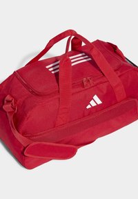 Bolsa de deporte roja fabricada en tejido texturizado, con asas dobles y una correa de hombro ajustable. Tres franjas blancas y detalle de logo.