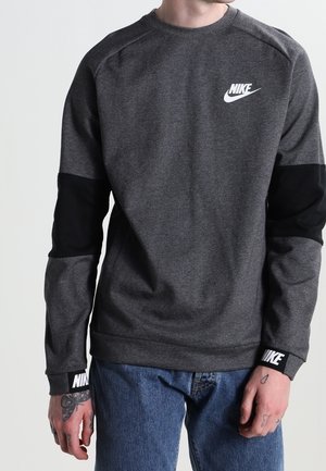 Mann trägt einen dunkelgrauen Nike-Sweatshirt mit schwarzen Ellbogenflicken, weißen Nike-Logos auf Brust und Bündchen, kombiniert mit blauen Jeans.