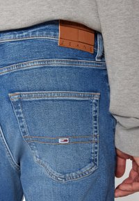 Modré džíny vyrobené z denimu s hnědou koženou nášivkou s nápisem "TOMMY JEANS." Mají šité zadní kapsy a texturovaný povrch.
