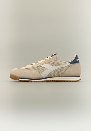 Beige und marineblaue Sneaker aus Mesh und Wildleder mit weißem Logo, Schnürung vorne und Kautschuksohle vor neutralem Hintergrund.