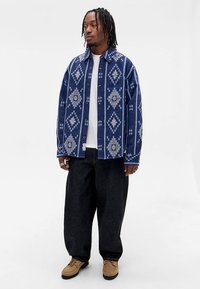 Camisa oversized azul com padrão geométrico, camiseta branca por baixo, calças largas escuras e botas castanho claro. Textura de mistura de algodão.