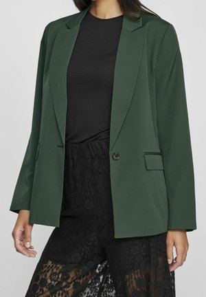 Vrouw die een donkerblauwgroene blazer draagt over een zwarte top en zwarte broek met kantpatroon, staand tegen een effen achtergrond.