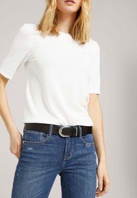 Top blanco de manga corta combinado con pantalones vaqueros de mezclilla azul y un cinturón negro con hebilla de plata. Los pantalones tienen detalles desgastados y bolsillos.