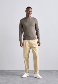 Brun genser med rund hals, beige plissert bukse og hvite joggesko. Genseren har en glatt tekstur, buksen har en avslappet passform.