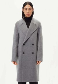 Grå ull dubbelknäppt kappa med stora slag, två framfickor och svarta knappar, uppvisad på en modell mot en vit bakgrund.