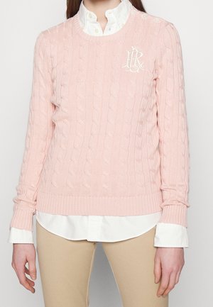 Femme portant un pull en maille torsadée rose pâle sur une chemise blanche à col avec un pantalon beige, montrée des épaules jusqu'aux hanches.