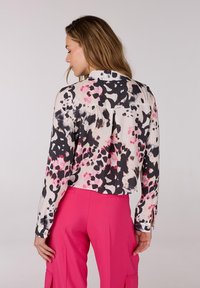 Korte blouse van lichte stof met een zwart, roze en crèmekleurig abstract patroon. Bevat een kraag en lange mouwen met knoopsluiten.