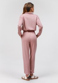 Ensemble de survêtement rose en deux pièces, comprenant un haut à col et un pantalon taille élastique. Tissu lisse avec des chevilles fuselées, porté avec des baskets blanches.