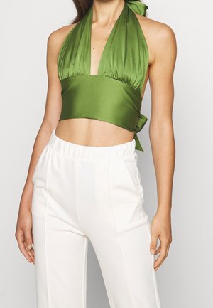 Kobieta ubrana w zielony, bezrękawnikowy krótki top z głębokim dekoltem w kształcie litery V oraz białe spodnie z wysokim stanem, stojąca na jednolitym tle.