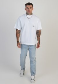 Camiseta blanca de manga corta con cuello redondo y detalle bordado, combinada con vaqueros azul claro y zapatillas gris y blanca.