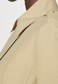 Trench-coat beige avec un col structuré, des manches longues et une texture lisse. Présente des coutures visibles et un détail de bouton sur le côté.