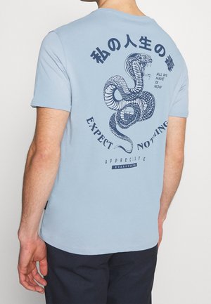 Lichtblauw T-shirt met een donkerblauw slangenmotief op de achterkant, omringd door tekstuele elementen. Korte mouwen en ronde hals.