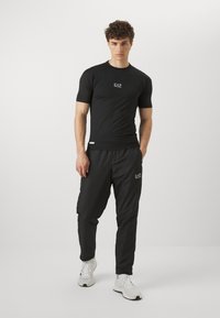 Svart atletisk t-skjorte med korte ermer og logo på brystet; kombinert med svarte smale joggebukser; begge laget av lett, pustende materiale.