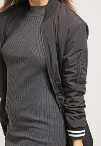 Giacca bomber nera con texture a costine, cerniera e tasca con zip sulla manica. Presenta polsini e colletto a costine a righe.
