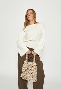 Biely top s dlhým rukávom a zvonovými manžetami, skombinovaný s hnedými nohavicami s širokými nohavicami a tkanou béžovou kabelkou na rameno s dekoračnými akcentmi.