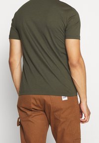 Olivgrünes T-Shirt mit kurzen Ärmeln und glatter Textur. Braune Cargo-Hose mit Seitentaschen und einem Markenpatch nahe der Taille.