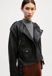 Casaco de motoqueiro de couro preto com um design encurtado, frente com fecho de correr, bolsos laterais e detalhes de botão. Combinado com um suéter de gola alta cinza por baixo.