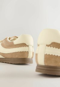 Brūnas un krēmkrāsas sporta kurpes ar zamšādas augšdaļu, fakturētām rakstām un gumijas zolēm. Dizainā ietvertas vilnainas detaļas sānos.