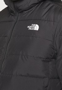 Musta puhvijakku vaakasuorilla tikatuilla osuuksilla, eteenpäin avattava vetoketju ja valkoinen logo vasemmalla ylhäällä. Sileä, kiiltävä kangas.