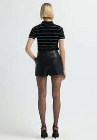 Donna bionda che indossa una maglietta a righe bianche e nere a maniche corte, pantaloni corti in pelle nera, collant neri trasparenti e tacchi alti neri, in piedi di spalle.