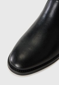 Botín negro de cuero con textura suave, punta redonda y una suela de goma duradera con detalles de costura a lo largo del borde.