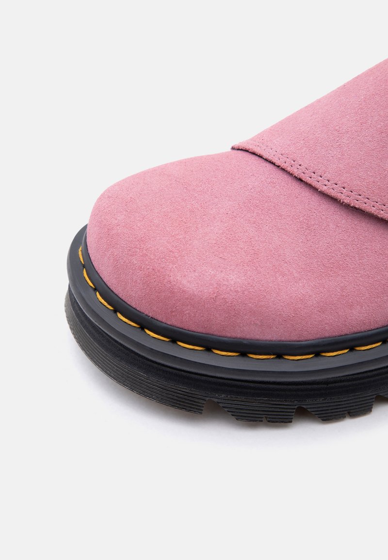 Primo piano della punta di una scarpa in camoscio rosa con suola spessa nera e cuciture gialle su sfondo bianco.