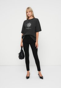 Overmaat zwart graphic t-shirt met witte print, gecombineerd met strakke zwarte broek. Accessoires zijn een gestructureerde zwarte tas en spitse zwarte hakken.