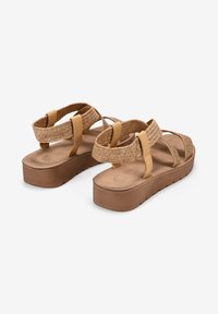 Sandalias de plataforma marrones con diseño de tiras trenzadas, suela plana y correas en el tobillo ajustables. Fabricadas con materiales sintéticos con acabado texturizado.