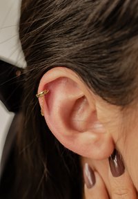 Goldene Creolen mit einem kleinen Kettendetail, die am oberen Teil des Ohrs befestigt sind. Die Haut des Ohrs wirkt glatt, mit sichtbarem dunklem Haar.