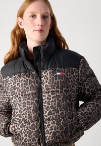 Pusset jakke med leopardmønster og svart nylon på toppen, høy krage, glidelås-lukking og logo-patch på brystet. Har ribbede mansjetter.