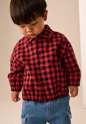 Unge barn iført en rød og sort ternet jakke og blå jeans, kigger ned med hænderne afslappet ned langs siderne.
